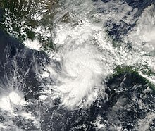 A photograph of a tropical storm very near the Pacific coast of Mexico; some of the clouds are spreading over the terrain, though most of it is in some closely grouped rainbands spiraling out to the south of the storm's center. Near the center is a slight dimple in the cloud tops, representing a primitive eye feature.