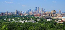 The Minneapolis skyline rises to its highest point at the center of the image, with the three tallest buildings standing out against a clear blue sky. Before the skyline are trees, university buildings, and residential complexes.