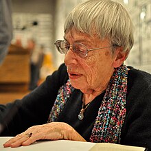 une femme âgée en dédicace avec lunettes, cheveux gris courts, une écharpe