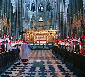 The choir master walking down the aisle of the abbey, with choirboys in red and white robes standing in stalls down its length
