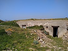 Photographie en couleur montrant un bâtiment en béton armé très épais, avec une porte à droite, au milieu d'une motte de terre et de l'herbe par dessus.