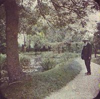 A grainy photo of a bearded man standing on a path before a tree and pond