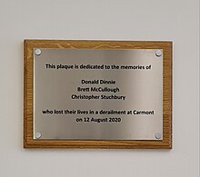 a simple plaque in brushed steel on a wooden backing, which reads "This plaque is dedicated to the memories of Donald Dinnie, Brett McCullough, Christopher Stuchbury who lost their lives in a derailment at Carmont on 12 August 2020"