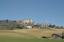 photo couleur d'un village perché au pied duquel s'étend une colline cultivée en pente douce.