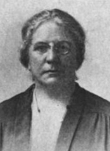 A middle-aged white woman with greying hair, wearing glasses and a dark suit jacket over a light-colored blouse