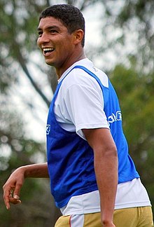 A smiling man with short and dark hair wears a white T-shirt under a blue training vest and pale yellow pants.