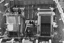A cavernous steel room, with two large rectangular boilers inside; a number of men are working on the machines.