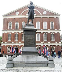 A statue on a pedestal of a man standing with his arms crossed. An inscription on the pedestal reads, "Samuel Adams, 1722–1803. A Patriot. He organized the Revolution and signed the Declaration of Independence." Behind the statue is a three-story brick building with many windows.