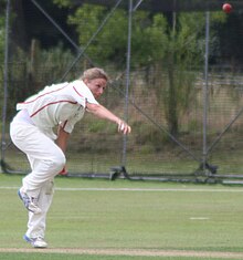 A woman with light brown hair wearing white cricket uniform with red piping has just bowled a cricket ball, still visible at the top right. She is bent over with her right arm extended in front of her. Cricket training nets are visible in the background.