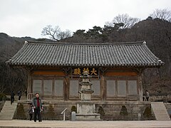 Photographie de l'extérieur du bâtiment d'un temple. Il est derrière une statue, et trois grands caractères chinois surplombent une porte.