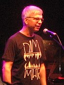 A gray-haired man with glasses and a black shirt standing in front of a microphone