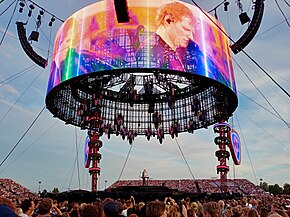 An outdoor stadium filled with spectators on the podiums and on the ground. In the middle Ed Sheeran performs on a raised stage, with video screens above him.