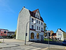 einzelnes Haus einer ehemaligen Häuserzeile, einspurig mit Zebrastreifen, im Hintergrund Bushaltestelle und Reklame