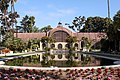 Photograph of the Botanic Building, one of several buildings in Balboa Park. A red conservatory building, it is fronted by a reflecting pool and surrounded by multiple species of trees.