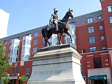 Harrison porte un bicorne et un uniforme militaire. La statue équestre en bronze se trouve un piédestal blanc devant des bâtiments en briques rouges.