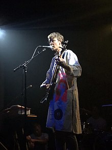 A person with glasses and short hair sings into a microphone onstage while playing guitar. She is wearing a long coat (akin to a lab coat) with several images painted on it, including a record and a pink cartoon of Cher with the slogan "What Would Cher Do?"