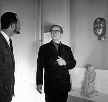 Image en noir et blanc d'un homme se tenant devant des objets d'art accrochés au mur pendant qu'un autre homme l'interroge.