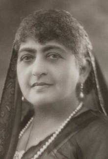 A middle-aged South Asian woman with greying hair, wearing a dark scarf over her head and a strand of pearls
