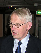 Half-portrait of a man in his seventies looking away from the viewer. He wears a suit and glasses.