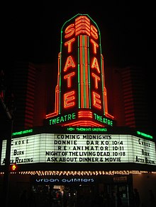 The neon marquee of a theater lists several notable cult films including Donnie Darko, Reanimator, and Night of the Living Dead.