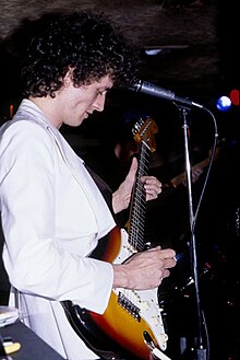 Man with curly dark hair wearing white jacket playing electric guitar standing close to a microphone