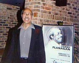 un homme Noir, cheveux gris et lunettes qui pose en souriant devant une affiche