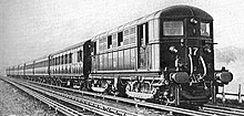 A black and white image of an electric locomotive hauling at least 6 coaches, shown with the electric locomotive on the right. A track in the foreground is electrified with the fourth rail system. The locomotive is shown with two pick-up shoes.