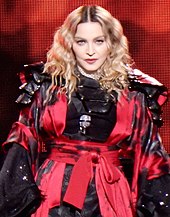 A blonde woman wearing a black dress with a red armor-like jacket and a microphone on her bosom.