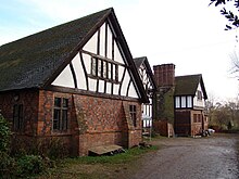 A house with three gabled bays seen from an angle. The outer bays have lower storeys in brick with a timber-framed gable. The middle gable is almost completely timber-framed on a low brick plinth. Between the second and third bays is a tall chimney stack.