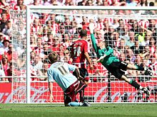 A football player in a red and white shirt and a football player in a light blue shirt are watching the ball approaching the goal, while the goalkeeper is diving to his right-hand side