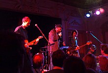 Three people performing on stage at Bush Hall, in the United Kingdom. They are illuminated by red and blue lights.