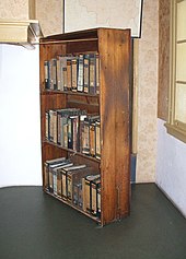 A three-shelf timber bookcase, filled with books, stands at an angle in front of a doorway to the Secret Annexe