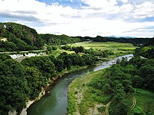River moving left to right in the mid ground surrounded by green grass and trees and mountains in the background