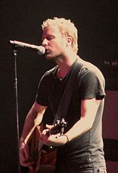 A fair-haired man singing into a microphone and playing a guitar