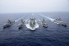 5 ships sailing in formation, center ship refuelling two ships, one each to the right and left; two additional ships visible in the background