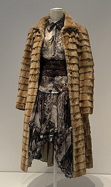 A light brown fur coat worn over a blouse and skirt in a fabric printed with birds, moths, and skulls