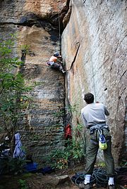 Grimpeuse faisant l'ascension d'un dièdre, assurée en moulinette par autre grimpeur positionné debout au pied de la voie d'escalade