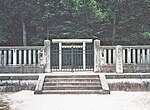 White torii gate behind a concrete fence in front of woods.