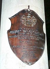 A white, cylindrical stone is marked with a copper-colored shield marking the burial place of Van Rensselaer, which includes Dutch text.