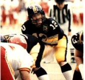 American football quarterback, Terry Bradshaw, prepares to receive a snap. He is wearing a black Pittsburgh Steelers jersey with a white number "12" and gold pants.