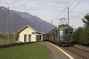 Single-story building with gabled roof next to side platform with green train on the tracks