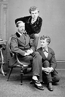 A studio, black and white photograph of three men, one is seated on an armchair, another is sitting on the floor beside him. The third is sitting behind them on a high stool and looks into the camera.