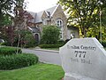 A close up picture of an address rock reading Hamilton Cemetery, 777 York Blvd.. The property has various bushes and trees and a large brick house in view.