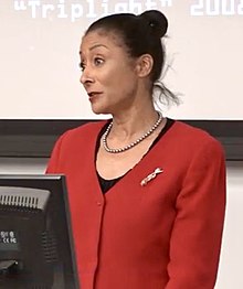 A woman with dark hair in a bun, wearing a red cardigan, there is the edge of the back of a computer screen in the forground and a part of a slide projection in the background.