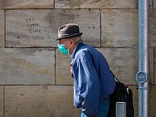 Un homme, avec chapeau, veste bleu, masque lui couvrant la bouche et le nez. Derrière sur un mur un grafiti : « It's Corona time » (C'est le temps du Corona).