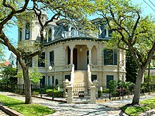 A Victorian-style home with a white exterior and a blue roof sits on a street corner with small trees partially obscuring the view. The front door is raised from street level with a small staircase leading up to the porch.