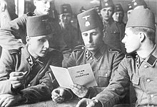 three soldiers in SS uniform and wearing fez headgear reading a pamphlet