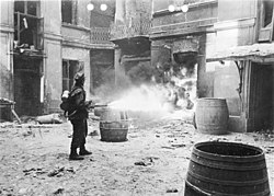 A black and white photograph of a German Verbrennungskommando soldiers using a flamethrower to set fire to ruins of a Warsaw building.