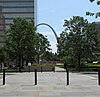 Four black bollards in the foreground, greenery in the center, and buildings in the background, with a tall grey arch partially visible in the distance