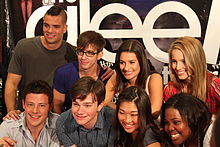 Cast of Glee huddled together, with the backdrop displaying the word "Glee" in white small fonts.